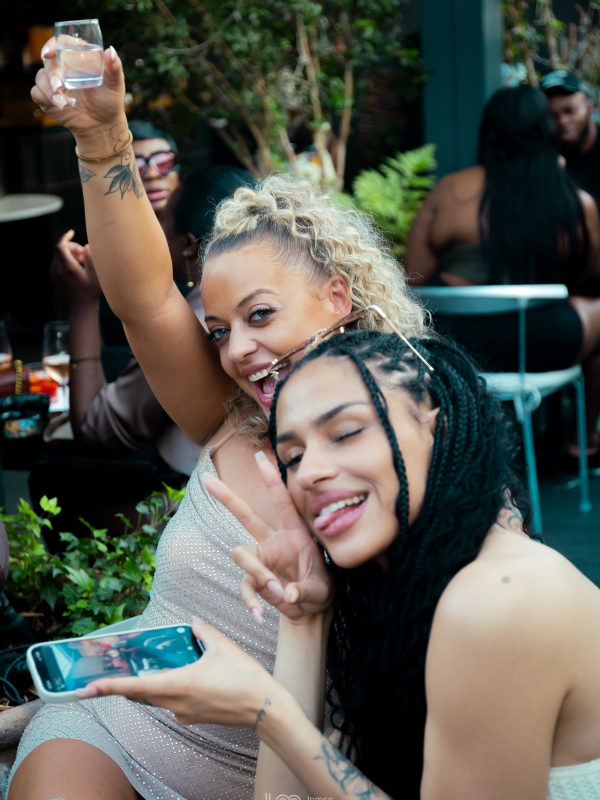 Two Women Smiling And Celebrating At An Outdoor Restaurant, One Holding Up A Glass In A Toast.